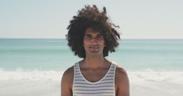 This image shows a young man with curly hair standing on a beach with the blue sea in the background. He is wearing a casual tank top and appears to be enjoying the sunny weather, making it ideal for use in travel brochures, summer vacation advertisements or promoting beachwear.