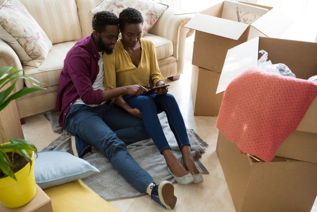 Couple sitting on floor surrounded by moving boxes, using digital tablet. Ideal for themes of moving, new home, technology in everyday life, and couple activities. Perfect for real estate, home decor, and lifestyle blogs.