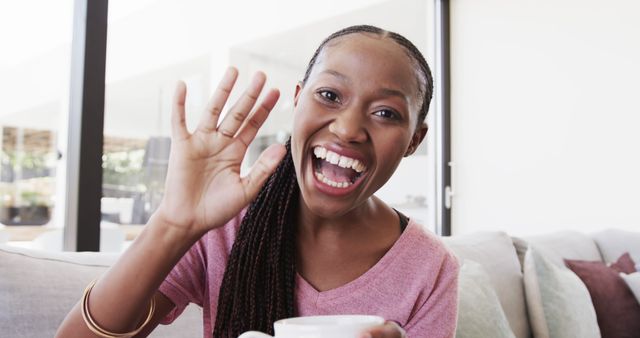 Cheerful African woman is waving and smiling, holding coffee mug, sitting comfortably on couch at home. Perfect for content related to home lifestyle, casual living, greetings, communication, morning routines, promotions for coffee or casual wear.
