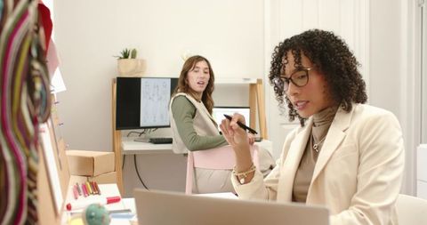 Mid-Adult Women Designers Collaborating in Bright Textile Studio Using Laptop and Stylus