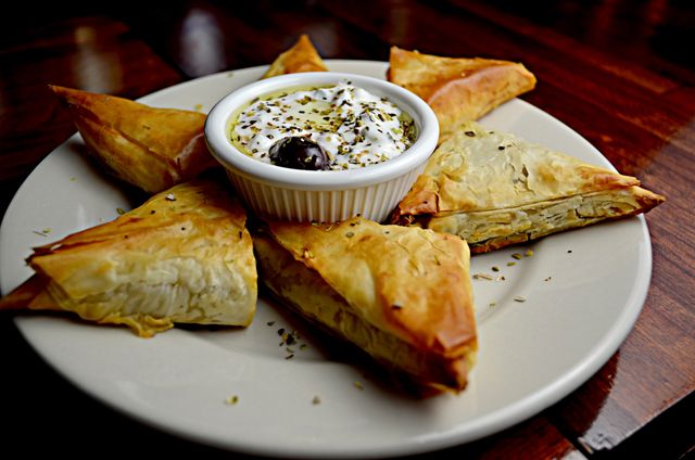 This image shows Spanakopita triangles arranged around a dip on a white plate, positioned on a wooden table. These phyllo dough pastries make an attractive appetizer, ideal for advertising Greek cuisine, restaurant menus, cooking blogs, or recipe websites.