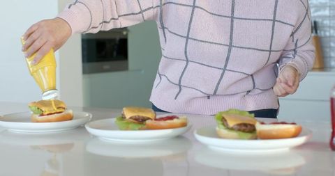 Woman squeezing mustard on three open-faced burgers at modern glossy kitchen counter