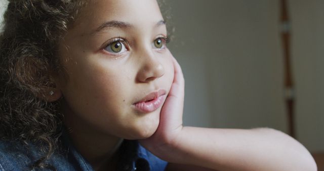 Young girl with curly hair contemplating with head resting on hand indoors. Useful for illustrating childhood innocence, moments of reflection, or emotional introspection. Ideal for educational materials, storytelling, and mental health awareness campaigns.