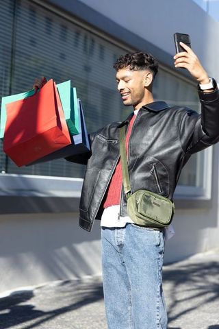 Indian man taking selfie with colorful shopping bags on urban sidewalk — street style