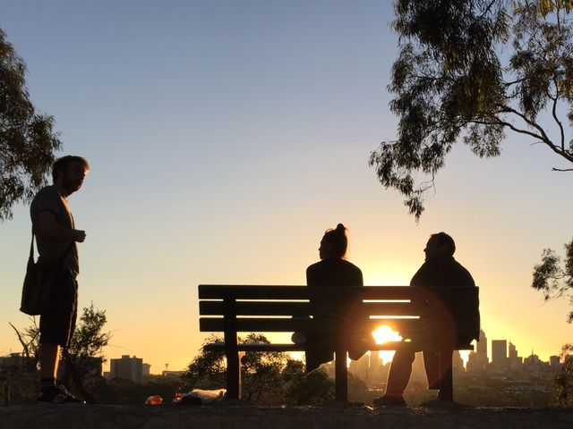Picture depicts a group of three friends, two seated on a bench, watching a sunset over a cityscape. Standing person gazes towards the setting sun. Suitable for content about friendship, outdoor activities, relaxation, urban nature spots, and city-inspired travel articles.