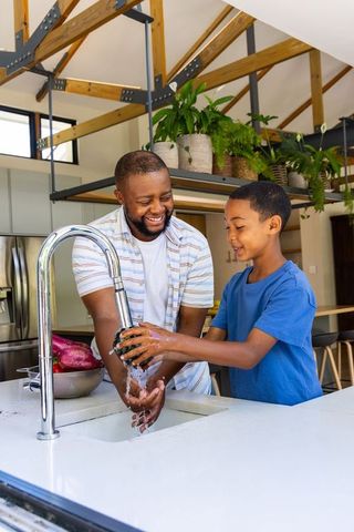 African american father and son washing produce at modern kitchen island, sharing smile