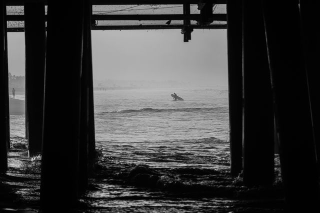 Silhouette of surfer catching a wave viewed from beneath a pier in black and white. Calm ocean creating contrast with dark framing of the pier. Perfect for use in adventure and surfing publications, travel brochures, or coastal lifestyle collections illustrating serene yet adventurous beach experiences.