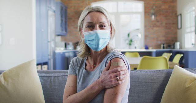 Portrait of happy senior caucasian woman wearing face mask in living room with bandage on her arm. senior health and lifestyle during covid 19 pandemic.