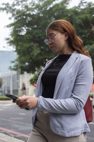 Young african american professional woman checking smartphone messages on wet city sidewalk