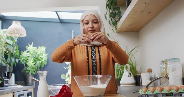 Image of smiling biracial woman in hijab baking in kitchen at home, breaking egg into mixing bowl. Happiness, health, inclusivity and domestic life.