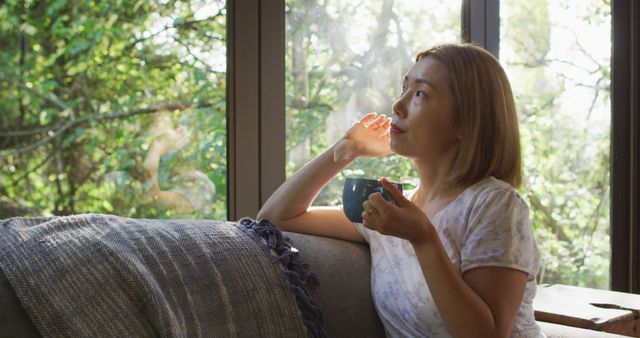 Asian woman looking through window and lying on couch with mug of coffee - Download Free Stock Photos Pikwizard.com