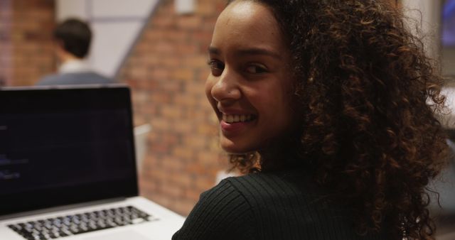 Young woman with curly hair smiling while working on laptop in an office setting. Ideal for depicting modern work environments, female professionals, or technology use in everyday life.