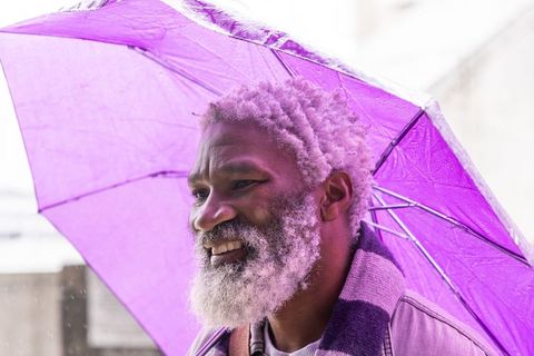 Senior african american man smiling under purple umbrella on rainy city street
