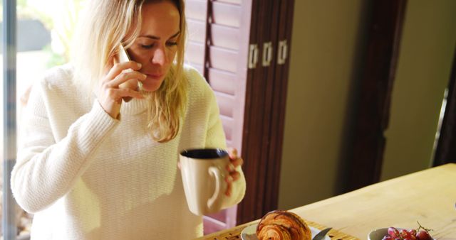 Woman sitting in kitchen, holding mug and smartphone, talking while enjoying morning breakfast with croissant and grapes. Useful for illustrating concepts of multitasking, relaxed mornings, home routines, and daily life. Can be used for lifestyle blogs, social media posts, and advertisements related to food, home living, and technology.