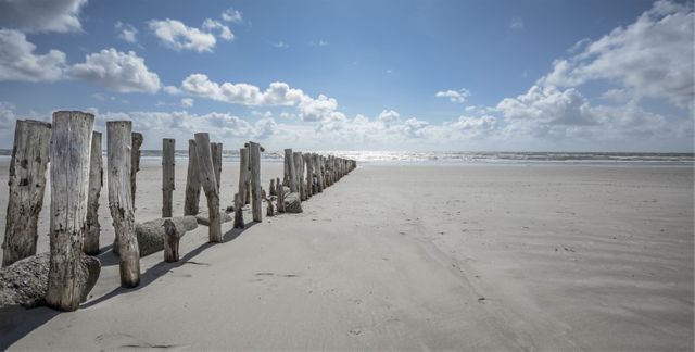 Wooden posts create a rustic barrier along picturesque ocean beach under bright blue sky with scattered clouds. Ideal for travel advertisements, nature blogs, wallpaper, backgrounds, promotional materials for beach resorts.