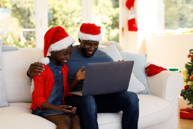 African-American father and daughter sitting on couch wearing Santa hats, looking at laptop. Christmas stockings hanging on wall in background. Perfect for holiday-themed promotions, family bonding, and technology use during festive season.