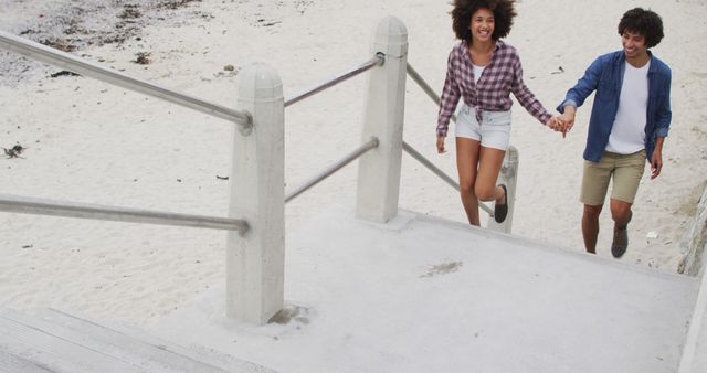 Captured in a lively and joyous moment, this happy couple is walking up stairs from a beach. Perfect for use in promotional materials for travel agencies, vacation packages, relationship blogs, and lifestyle magazines. The casual clothing and bright scenery are ideal for conveying adventure, relaxation, and romantic getaways.