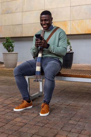 African american man smiling and texting on smartphone while sitting on bench in plaza
