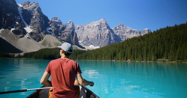 Young man enjoying serene kayaking experience on clear mountain lake, surrounded by lush forest and towering peaks. Ideal for use in travel blogs, adventure tourism promotions, outdoor activity guides, nature documentaries, and relaxation-focused advertisements.