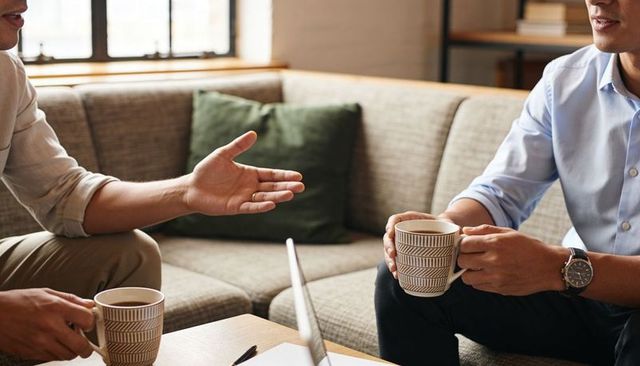 Colleagues chatting over coffee on sofa with laptop in cozy office lounge