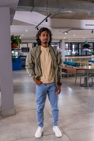 Indian man standing in modern coworking lounge wearing olive bomber jacket and white sneakers