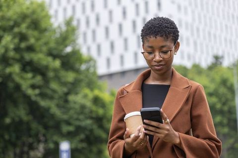 African american woman checking smartphone while holding to-go coffee cup on city street