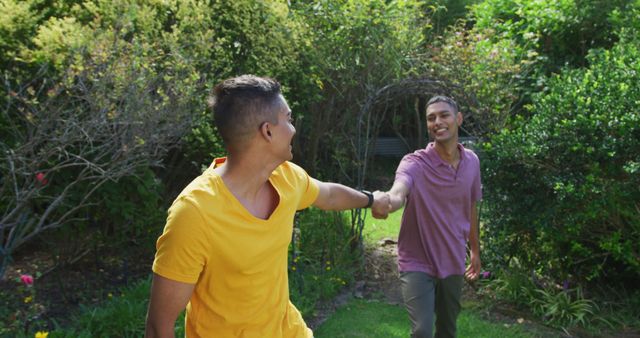 Smiling biracial gay male couple holding hands walking in garden. staying at home in isolation during quarantine lockdown.