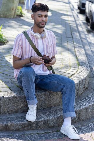 Asian man sitting on curb holding snack, wearing headphones and green crossbody bag