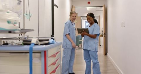 Smiling nurses collaborating on tablet in hospital corridor beside medical supply cart