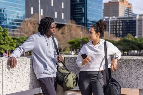 Urban friends leaning on waterfront railing, chatting in gymwear with duffel bags and smartwatch