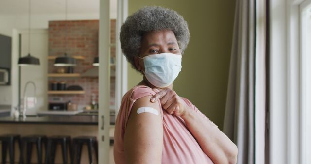 Senior woman standing indoors showing vaccination bandage on arm proudly. Wears face mask for safety. Ideal for use in health and wellness campaigns, vaccination promotion, COVID-19 pandemic awareness, and articles on senior care and health policies.