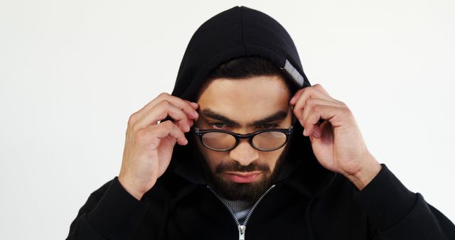 Man adjusting glasses while wearing black hoodie. White background emphasizes he is serious and focused. Useful for advertisement, marketing campaigns, or articles focusing on fashion, style, or concentration.