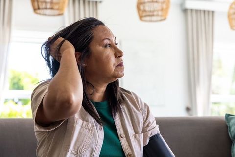 Mature woman sitting on sofa touching hair wearing beige overshirt and blood pressure cuff
