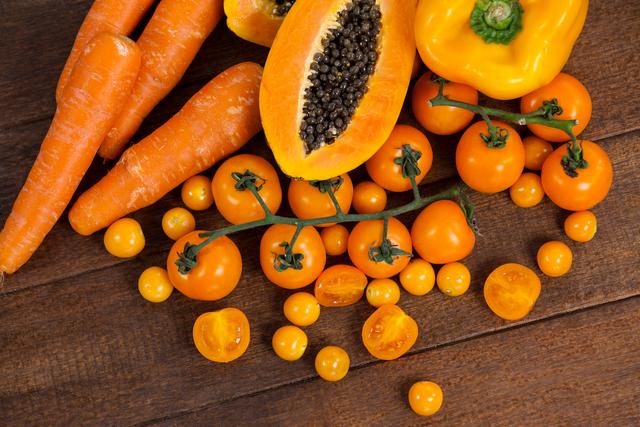 This vibrant image showcases a variety of fresh orange vegetables and fruit, including carrots, papaya, tomatoes, and a yellow bell pepper, arranged on a wooden table. Ideal for use in health and wellness blogs, diet and nutrition articles, vegan and vegetarian recipe websites, and organic food promotions.