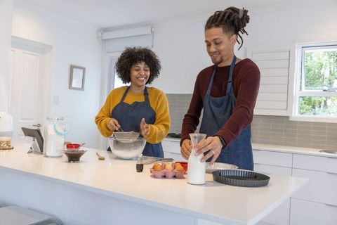 Happy couple baking together on bright kitchen island, whisking batter and pouring milk