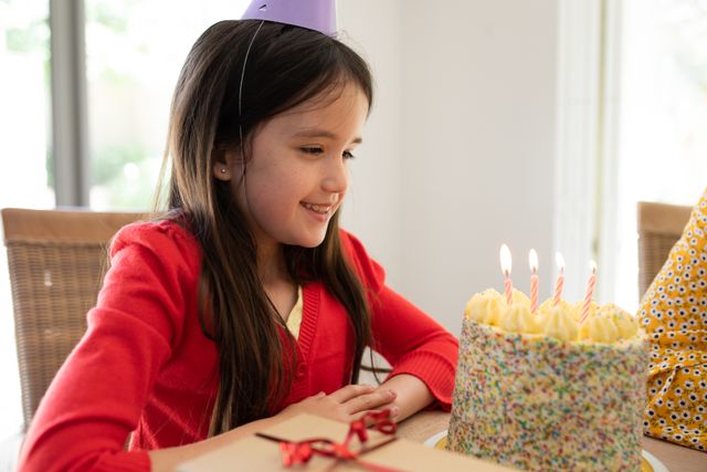 Little caucasian girl wearing a birthday hat sitting on the dinner table in front of a birthday cake with four candles. sitting beside her is her mom.