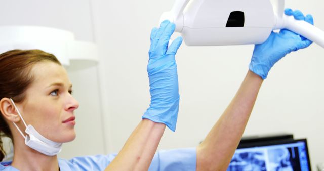 Female dentist wearing blue gloves and a mask, positioning an x-ray machine in a dental clinic. Ideal for use in articles related to dental healthcare, professional dentistry, and dental equipment. Can be used to illustrate topics on dental hygiene, dental check-ups, and healthcare settings.