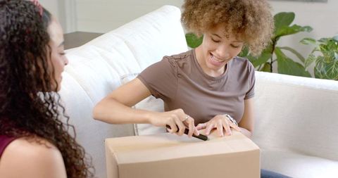 Smiling woman unboxing delivery on couch with friend using utility knife