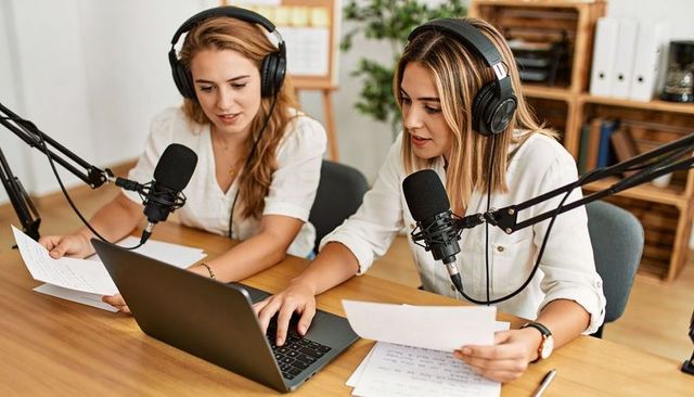 Two female podcasters recording together at wooden desk with laptop, notes and headsets