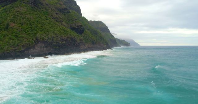 Majestic coastal cliffs rise above turquoise waters as waves crash against the shoreline in misty weather, featuring lush green vegetation. Ideal for use in travel blogs, nature magazines, and environmental conservation promotions.