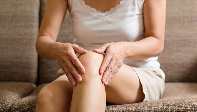 Middle-aged woman holding knee on couch experiencing joint pain seeking comfort at home