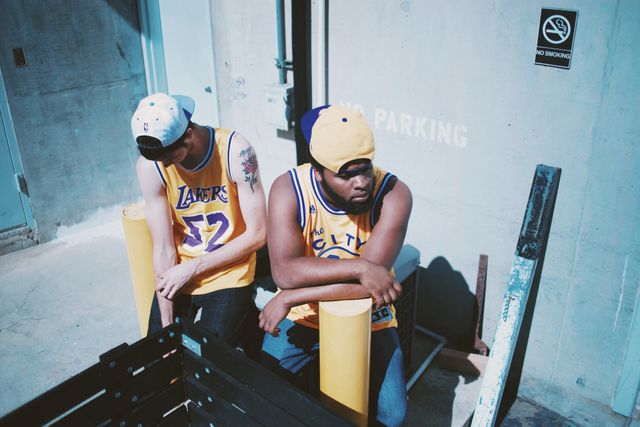 Two friends are sitting outdoors, dressed in Lakers basketball jerseys with caps. They appear relaxed and casually engaged, enjoying each other's company. The scene includes urban elements such as a no-smoking sign and some barriers, creating a contemporary, youthful vibe. This image is ideal for use in content related to sports, friend bonding, urban lifestyle, and casual summer activities.