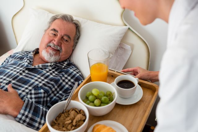 Senior man in bed receiving breakfast from a nurse in a nursing home. The tray includes a glass of orange juice, grapes, coffee, cereal, and a croissant. Ideal for use in healthcare, elderly care, and nursing home promotional materials, as well as articles on senior health and wellness.