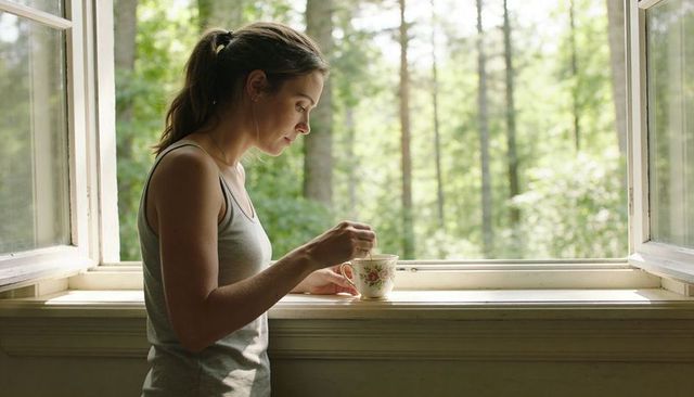 Woman stirring tea by open window holding floral cup overlooking lush forest morning calm