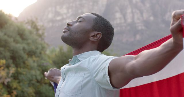 African American man extends arms with joy, draped in flag, surrounded by nature and mountains. Perfect for concepts of freedom, celebration, diversity, pride, national holidays, and outdoor activities.