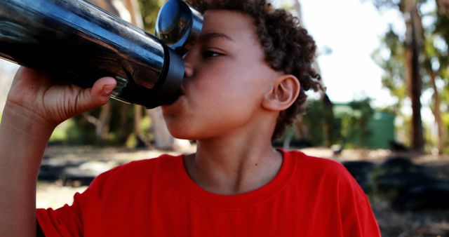 Young boy wearing a red shirt drinking from a water bottle while outdoors on a sunny day. The background features trees and natural elements, creating a fresh and healthy atmosphere. This image can be used for promoting hydration, healthy lifestyles for children, outdoor activities, or children's summer camps.