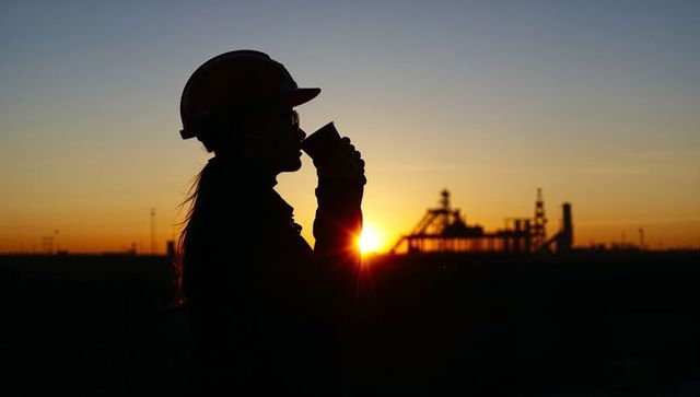 Silhouetted female worker sipping coffee at industrial sunrise, hard hat goggles