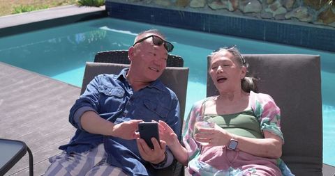 Senior asian couple relaxing by turquoise pool, sharing smartphone and drink on lounge chairs