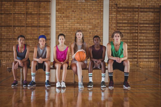 Group of high school basketball players sitting on a bench in a gymnasium. They are wearing sports uniforms and appear focused and ready for practice or a game. This image can be used for promoting youth sports programs, school athletics, teamwork, and fitness initiatives.