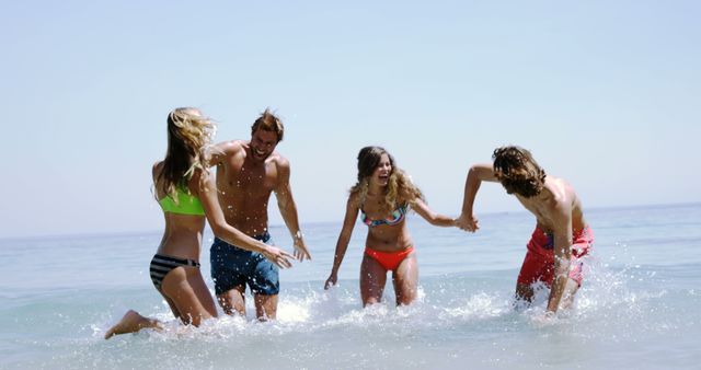Young adults are enjoying a sunny day at the beach, splashing in the shallow ocean water, with copy space. Their joyful expressions and active engagement with each other capture the essence of summer fun and friendship.